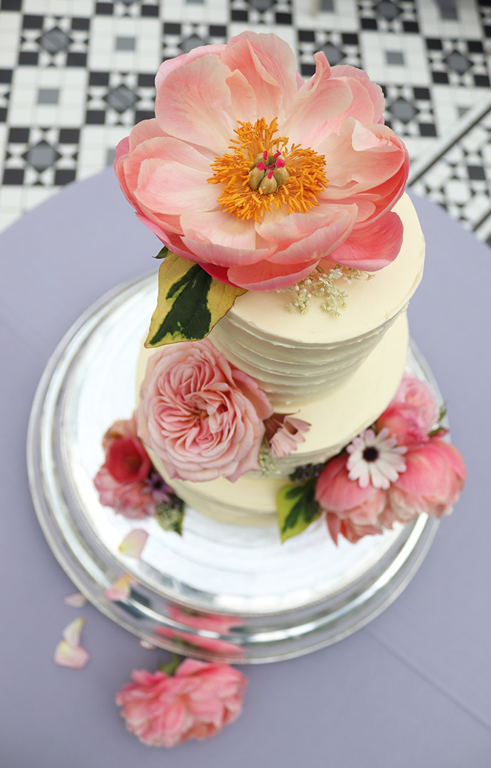 A wedding cake in the conservatory at Horniman Museum & Gardens. Three tiers of award-winning dairyfree Tunisian Orange & Almond Cake, filled and frosted with orange Swiss meringue buttercream. All ingredients gluten-free. Served for dessert with roast peaches. Decorated with organic edible peonies and roses from Maddocks Farm Organics. Price includes tasting consultation, and delivery and assembly at your London venue